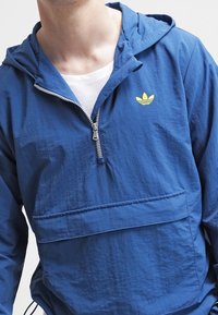 Modrá lehká bunda s kapucí, polovičním zipem vpředu, přední kapsou a žlutým logem na hrudi. Hladká textura, ležérní design.