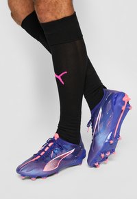 Des crampons de football violets et roses avec un design texturé, présentant des accents roses et une chaussette noire avec un logo rose.