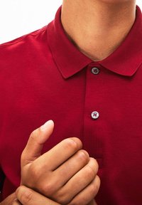 Czerwona koszulka polo wykonana z miękkiego materiału, z kołnierzykiem, dwu guzikową plisą z szarymi guzikami oraz małym detalem loga nad dołem.