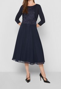 Mujer vestida con un vestido de encaje azul marino con una chaqueta corta a juego y zapatos de tacón alto negros de punta.