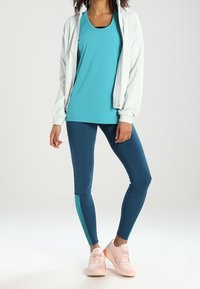 Turkos linne, vit zip-up-jacka, marinblå leggings med teal rand, och ljusrosa sneakers. Släta texturer och atletisk design.