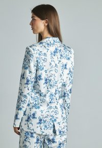 Completo floreale in tessuto bianco con motivi blu, caratterizzato da una vestibilità su misura, rever a punta e uno spacco singolo sul retro.