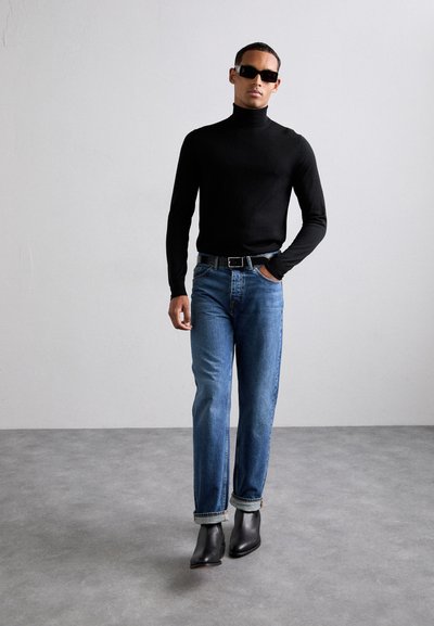 Svart polotröja, blå jeans med uppvikt fåll, svarta läderstövlar knappt ovanför ankeln och mörka solglasögon; minimalistisk design, figurnära siluett.