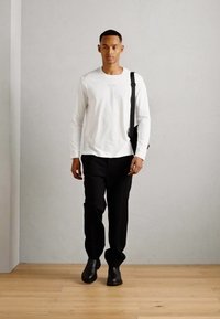 Cămașă albă cu mâneci lungi, pantaloni negri și cizme negre până la gleznă. Modelul poartă o geantă neagră. Design simplu, texturi netede și culori neutre.