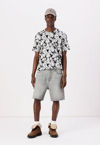 Sort og hvid blomstret t-shirt, grå denim shorts, brune støvler med pelsdetaljer, iført en lys grå kasket, stående på en ensfarvet baggrund.