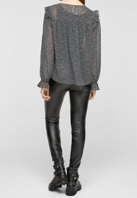 Blouse noire transparente avec des manches froncées et un motif floral subtil, associée à un pantalon en simili cuir noir moulant et des bottines noires robustes.