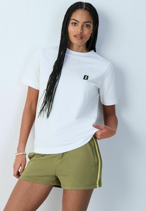 Kvinde med lange fletninger iført en hvid t-shirt med et lille sort logo og olivengrønne shorts med sidelommer, stående op ad en hvid væg.