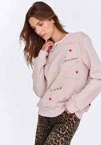 Světle růžová mikina s vícolorovými nápisy a grafikou srdcí; v kombinaci se džínami s leopardím potiskem, které mají žebrované manžety a lem.