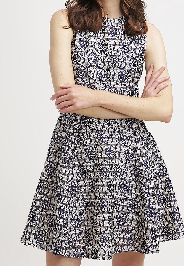 Navy- und cremefarbenes Spitzekleid mit engerem Oberteil und ausgestelltem Rock. Es besitzt ein ärmelloses Design und ein aufwendiges Blumenmuster.