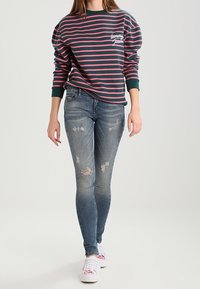 Jersey de rayas en azul marino y rojo, con un logo; vaqueros azules desgastados; zapatillas blancas con detalles florales. Detalle de un conjunto informal.