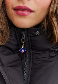 Zbliżenie na usta i podbródek kobiety w czarnej kurtce z niebiesko-różowym, wachlarzowatym suwakiem.