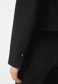 Zwarte op maat gemaakte blazer met een gestructureerde stof. Kenmerken zijn een slanke snit, knoopdetails op de mouw en een discrete zak op de achterkant.
