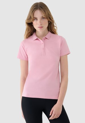 Młoda kobieta o falujących blond włosach, ubrana w jasnoróżową koszulkę polo z krótkim rękawem i czarne spodnie na jednolitym tle.