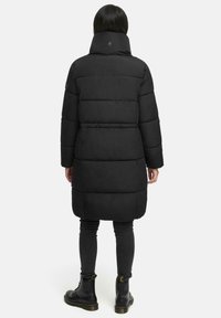 Black padded long coat with a high collar, featuring a quilted design and a logo detail at the back. Paired with dark slim jeans and boots.