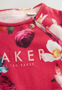 Czerwona koszula w kwiaty z różowymi i białymi wzorami, metalowe zapięcie na zatrzask oraz napis "BAKER by TED BAKER" wytłoczony na złoto.