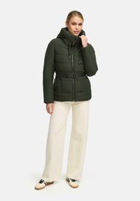 Giacca puffer verde imbottita con colletto alto, vita cintrata, chiusura con zip e cordini regolabili, abbinata a pantaloni a gamba larga beige chiaro.