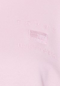 Vaaleanpunainen puuvillaneule, jossa on brodeerattu logo, jossa lukee brändin nimi "TOMMY HILFIGER", sekä suorakulmainen grafiikka keskellä.