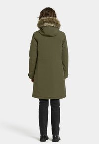 Olivengrøn parka med hætte foret med kunstpels, lange ærmer og en kant, der når midt på låret. Båret over sorte bukser og støvler.