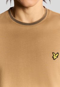 Sandfärgad t-shirt med ribbade svarta kanter och en svart och gul broderad fågologotyp på vänster bröstområde. Slät tygstruktur.