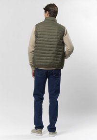 Bărbat purtând o vestă matlasată verde măsliniu, cămașă bej cu mâneci lungi, blugi albaștri și adidași bej, stând cu spatele spre camera foto pe fundal alb.