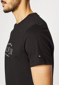 Camiseta de algodón negra con cuello redondo, mangas cortas y un pequeño acento bordado en rojo, blanco y azul en la manga izquierda.