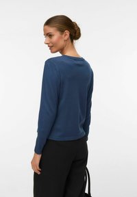 Femme avec les cheveux en chignon portant un pull bleu à manches longues et un pantalon noir, regardant par-dessus son épaule sur un fond blanc uni.