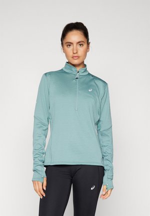 Haut de sport long en teal avec un col zippé mi-haut, un motif texturé et des poignets avec trou pour le pouce. Associé à des leggings noirs avec un logo.