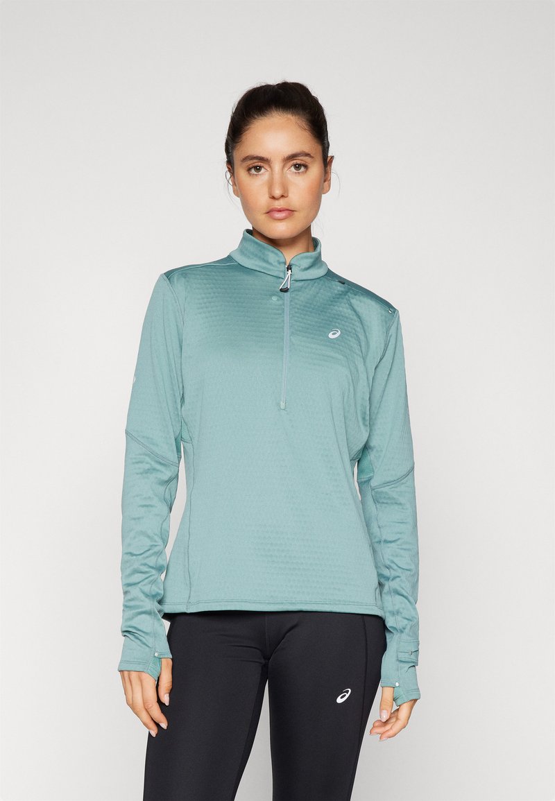 Teal sportieve longsleeve top met een halve ritssluiting, textuurpatroon en duimgaten in de mouwen. Gecombineerd met zwarte leggings met een logo.