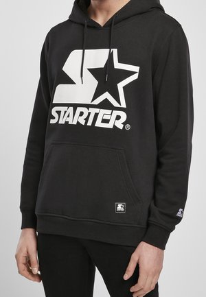 Sweat à capuche noir avec logo blanc « Starter » et étoile sur la poitrine, poche kangourou à l'avant et cordons sur la capuche porté par une personne cadrée au niveau du cou et des cuisses.