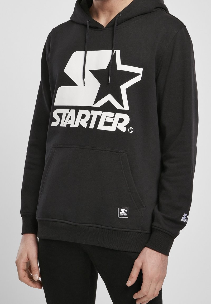 Sweat à capuche noir avec logo blanc « Starter » et étoile sur la poitrine, poche kangourou à l'avant et cordons sur la capuche porté par une personne cadrée au niveau du cou et des cuisses.