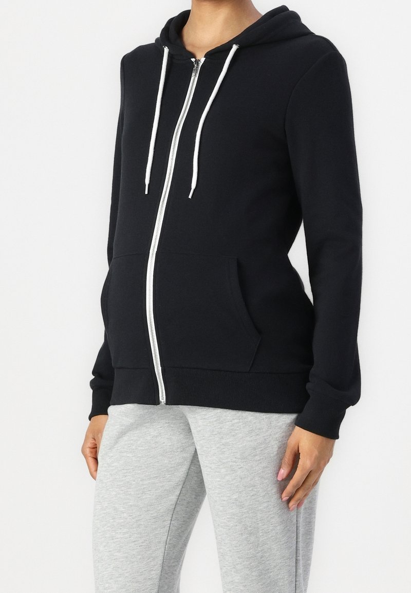 Hoodie noir à fermeture éclair avec cordons blancs, poches avant et ourlet côtelé. Associé à un pantalon de survêtement gris, doté d'une texture en coton doux.