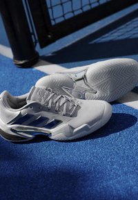 Biele športové topánky Adidas s modrými pásikmi ležiace na modrom tenisovom kurte blízko čierneho sieťového stĺpa a bielej ohraničujúcej čiary.