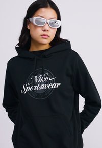 Sweatshirt preto com capuz e bolso frontal, apresentando um logótipo gráfico branco que diz "Nike Sportwear" no centro.