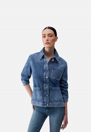 Mujer con chaqueta vaquera de manga remangada y bolsillo en el pecho con monograma y tachuelas, combinada con vaqueros a juego.