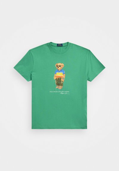 T-shirt en coton vert avec un motif représentant un ours portant une chemise bleue et jaune, avec le texte "POLO BEAR by RALPH LAUREN" en dessous.