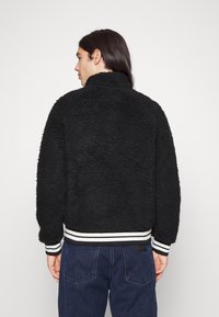 Sort, fluffy bomberjakke med høj krave, struktureret stof og ribbede manchetter og talje i hvid stribe. Har en afslappet pasform.