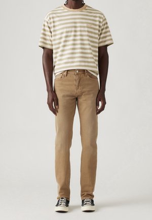 Mann trägt beiges gestreiftes Levi's T-Shirt, hellbraune Jeans und schwarz-weiße Sneakers und steht vor schlichtem Hintergrund.