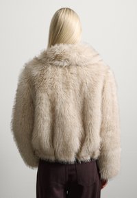 Giacca in pelliccia sintetica beige, caratterizzata da un colletto alto, una vestibilità larga e una texture morbida con una finitura lanosa. Sotto si indossano pantaloni marroni.