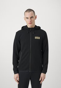 Felpa nera con zip, in tessuto texturizzato, cappuccio regolabile, tasche frontali e logo dorato sul petto. Polsini e orlo lisci.
