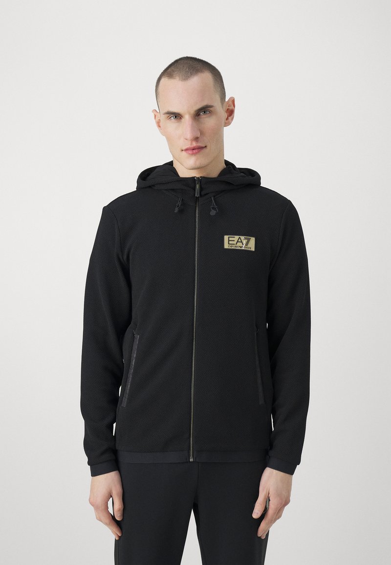 Felpa nera con zip, in tessuto texturizzato, cappuccio regolabile, tasche frontali e logo dorato sul petto. Polsini e orlo lisci.