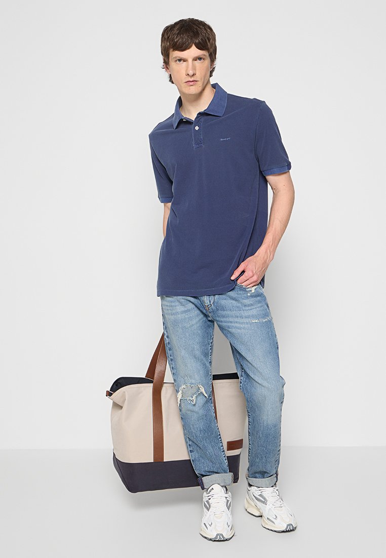 Mężczyzna w granatowej koszulce polo i przetartych dżinsach stoi z beżową i granatową dużą torbą na podłodze na tle białej ściany.