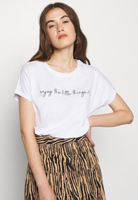 Tricou din bumbac alb, cu mâneci scurte și decolteu rotund. Prezintă un text cursiv negru "bucură-te de lucrurile mici" în față.