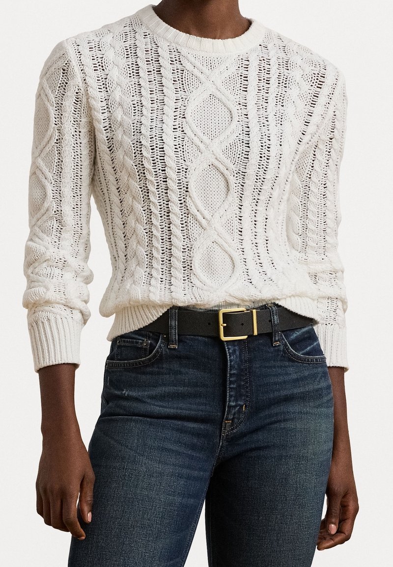 Personne portant un pull en maille torsadée crème rentré dans un jean bleu foncé avec une ceinture noire avec une boucle dorée.