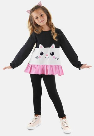 Schwarzes Langarmshirt mit einem weißen Katzenkopf-Design, pinkem Rüschen-Saum, kombiniert mit schwarzen Leggings und hellrosa Sneakers. Glitzernde Katzenohren.