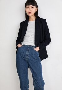 Marineblauwe textuur blazer over een grijs shirt, gecombineerd met hoge taille blauwe jeans. De blazer heeft een gestructureerd ontwerp en zichtbare stiksels.