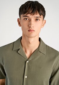 Młody mężczyzna z krótkimi ciemnymi włosami, ubrany w oliwkową koszulę z krótkim rękawem zapinaną na guziki, z małym haftowanym logo na piersi.