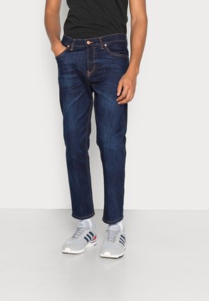 Homme portant un jean bleu foncé, une chemise noire et des baskets blanches avec des rayures noires, debout devant un fond uni.