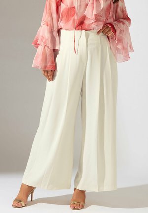 Kobieta w białych spodniach o szerokich nogawkach, przezroczystej różowej bluzce w kwiaty z falbanami na rękawach oraz złotych sandałach na obcasie, stojąca na jednolitym tle.