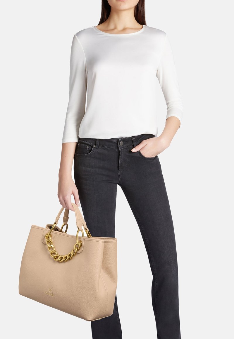 Borsa tote in pelle beige con finitura testurizzata, hardware in catena dorata e doppi manici. Il modello indossa una maglietta bianca e jeans scuri, con uno stile casual.