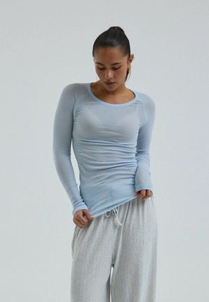 Frau schaut nach unten, trägt ein hellblaues, eng anliegendes Langarmshirt und lockere graue Hose mit Kordelzug, hält den Saum des Shirts.